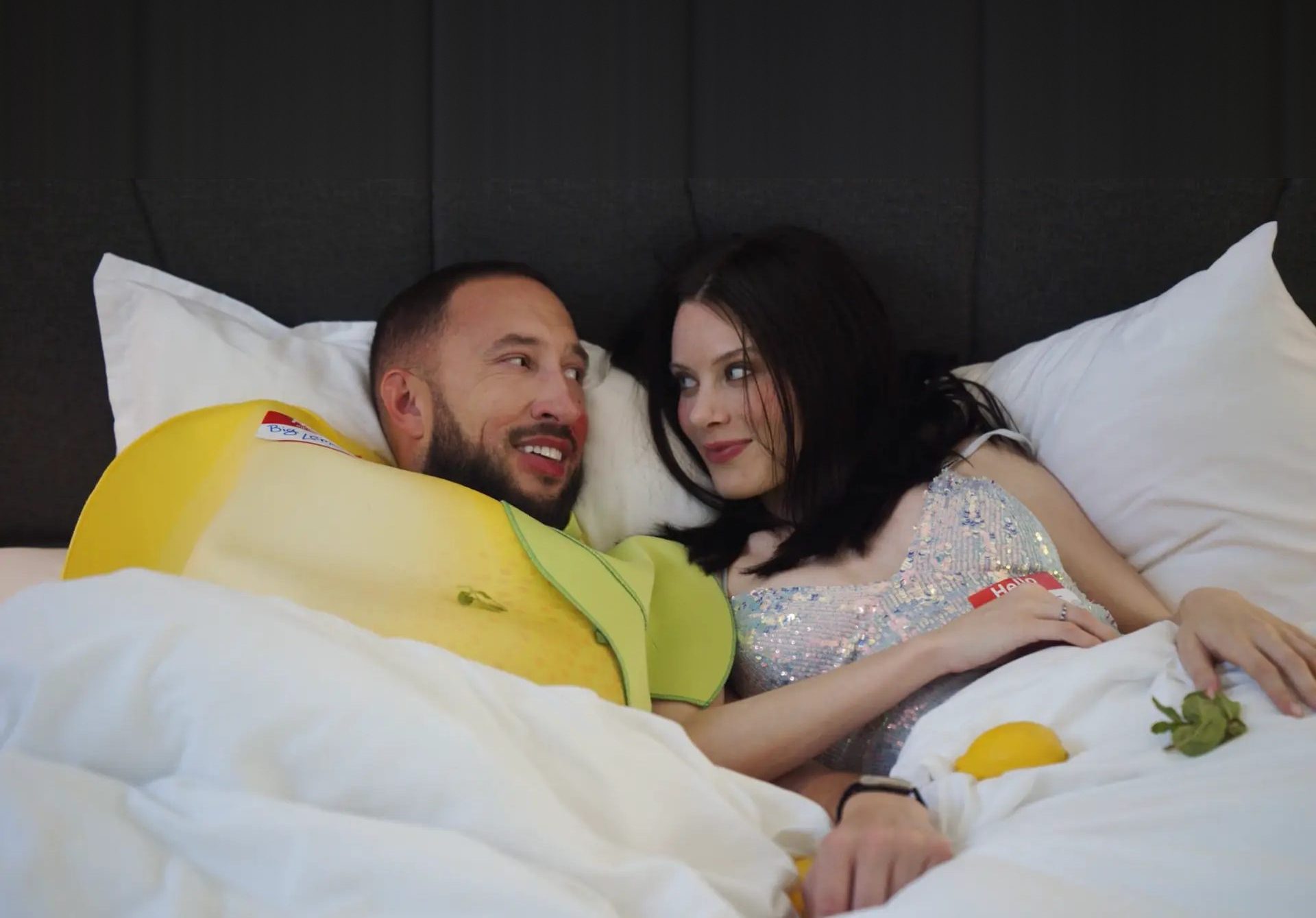 Two people lying in bed together, one in a yellow lemon costume with a name tag, the other in a sequined top