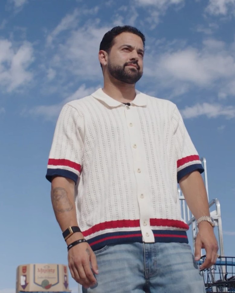 Man in a cream knit polo with red and navy striped cuffs and light-wash jeans, shot from a low angle against a bright blue sky, a ladder and stock carts behind him