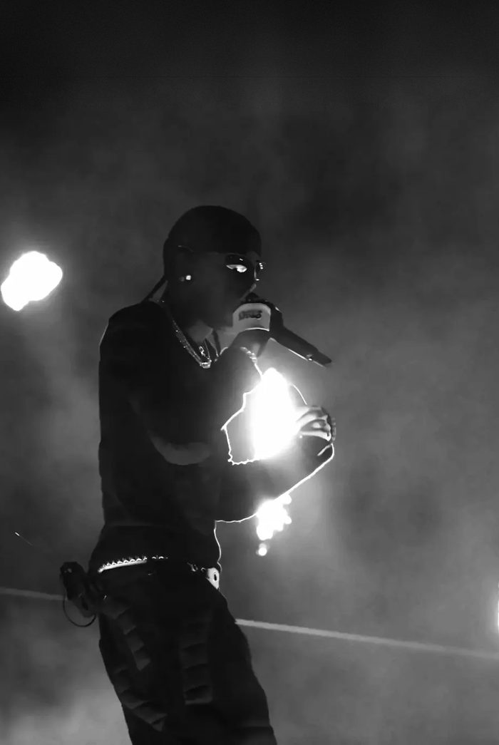 Black-and-white film still of a performer in profile holding a microphone to his mouth, backed by stage smoke and a blown-out light